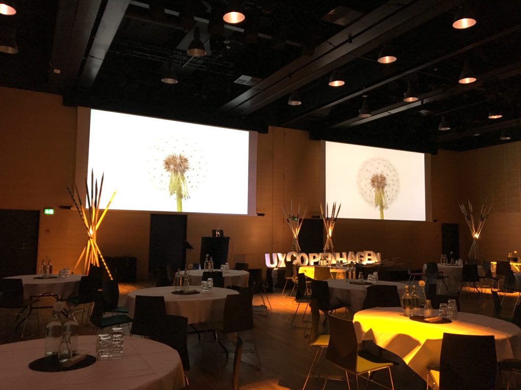 Day 1: Overall view of the auditorium. Screens shows a animation explaiting the origin of the UX Copenhagen logo